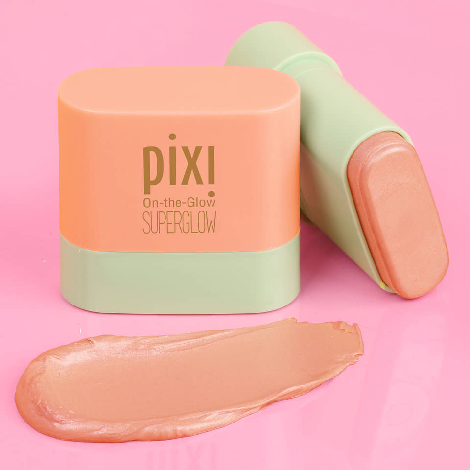 Pixi On-the-Glow SuperGlow stick blush shown with its peach-colored cap and pale green base, alongside a smear of the peachy-pink cream product on a solid pink background. view 1