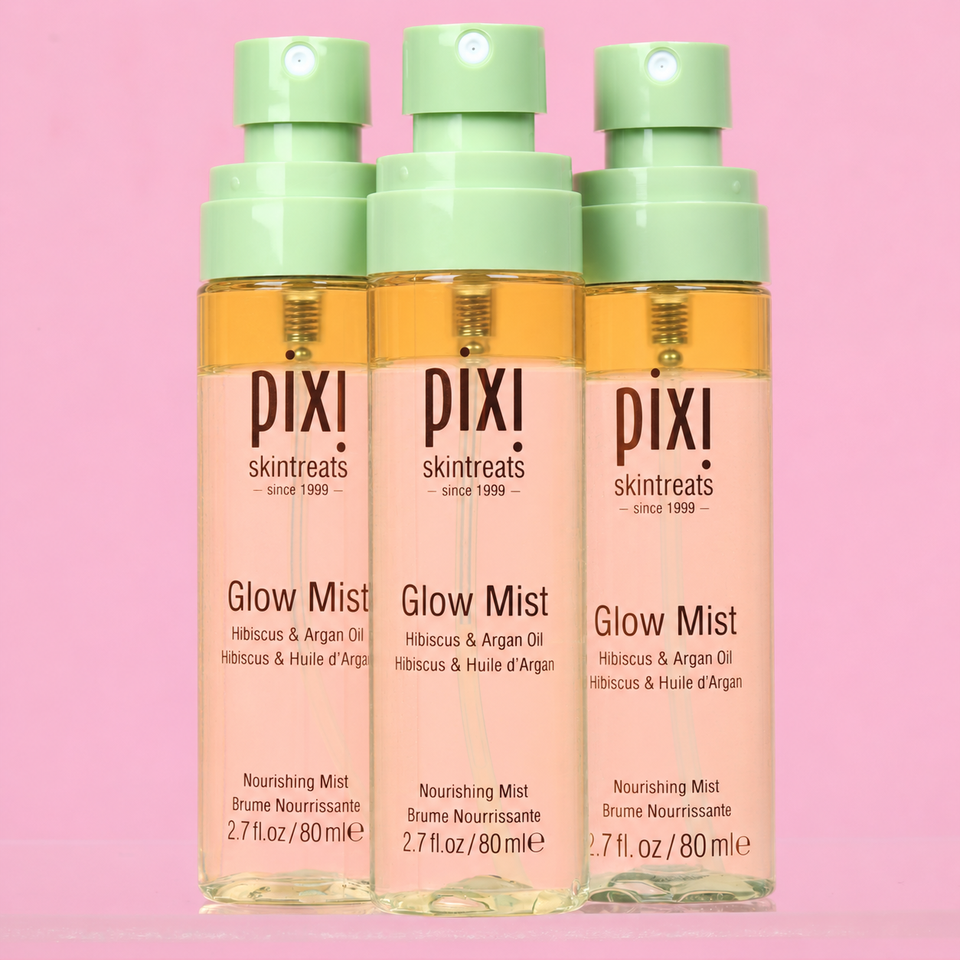 Three Pixi Skintreats Glow Mist bottles with pale green pump tops, filled with a warm golden liquid, set against a solid pink background. view 1