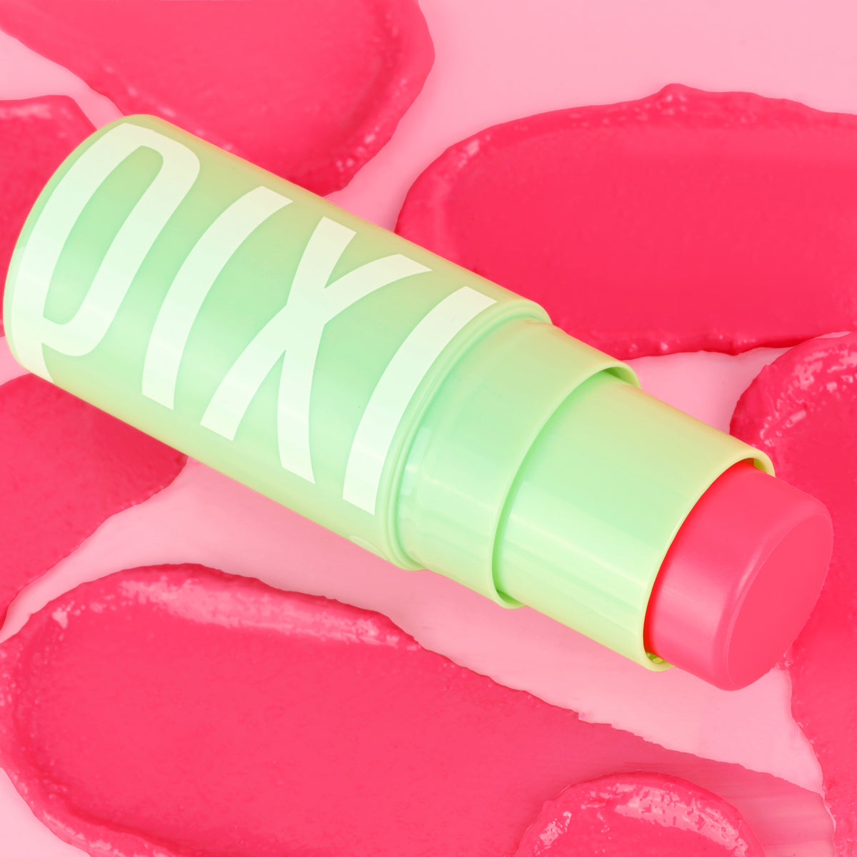 A close-up of a Pixi +Hydra LipTreats tube lying diagonally on a pink background with pink lip-shaped decorative elements. The tube is light green with large white Pixi lettering, and the balm stick itself is a bright pink.
